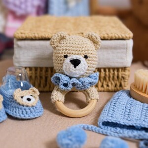K&ouml;nnte beinhalten: Ein geh&auml;keltes Baby-Set in Blau und Beige. Das Set beinhaltet eine Teddyb&auml;r-Rassel, ein Paar Booties, eine M&uuml;tze und eine B&uuml;rste.