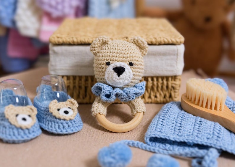K&ouml;nnte beinhalten: Ein geh&auml;keltes Baby-Set in Blau und Beige. Das Set beinhaltet eine Teddyb&auml;r-Rassel, ein Paar Booties, eine M&uuml;tze und eine B&uuml;rste.
