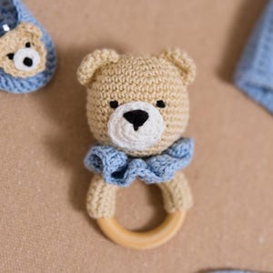 K&ouml;nnte beinhalten: Ein geh&auml;keltes Teddyb&auml;r-Rassel mit einem Holzring. Der B&auml;r ist beige mit einem blauen R&uuml;schenkragen und schwarzen Augen.