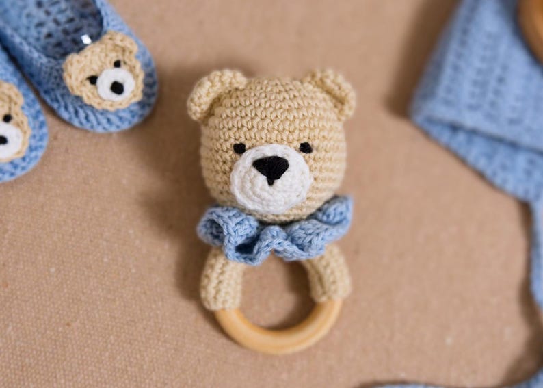 K&ouml;nnte beinhalten: Ein geh&auml;keltes Teddyb&auml;r-Rassel mit einem Holzring. Der B&auml;r ist beige mit einem blauen R&uuml;schenkragen und schwarzen Augen.
