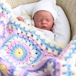 May include: A newborn baby sleeping in a white wicker basket, swaddled in a colourful crocheted blanket with pink, blue, yellow, and white colours. The baby is wearing a white hat.