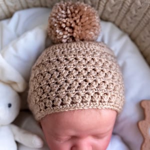 May include: A tan crocheted baby hat with a fluffy pom pom on top. The hat is worn by a baby sleeping on a white blanket.