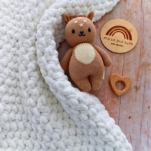 May include: A white chunky knit blanket, a brown knitted deer toy with a cream belly, a wooden heart-shaped teether, and a round wooden plaque with a rainbow graphic and the text Maisie and Ruth BABY CROCHET.