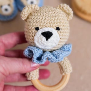 以下が含まれることがあります： 青いフリルと木製リングが付いた、かぎ針編みで作ったテディベアのおしゃぶり。おしゃぶりは手で持たれています。