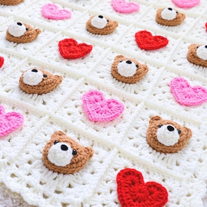 May include: A white crocheted blanket featuring a grid of square panels. Each panel contains either a small brown and white bear face applique or a heart applique in solid red or bright pink. The bear faces have black eyes and noses.
