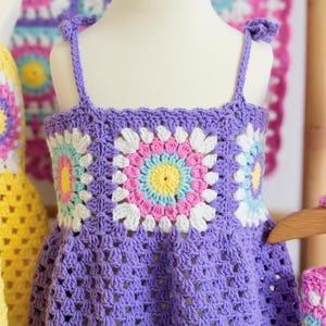 May include: A lilac-coloured, handmade crochet dress for a child, shown on a mannequin. The dress has a floral pattern on the bodice with white, pink, yellow, and blue details. The straps are tied at the shoulders.