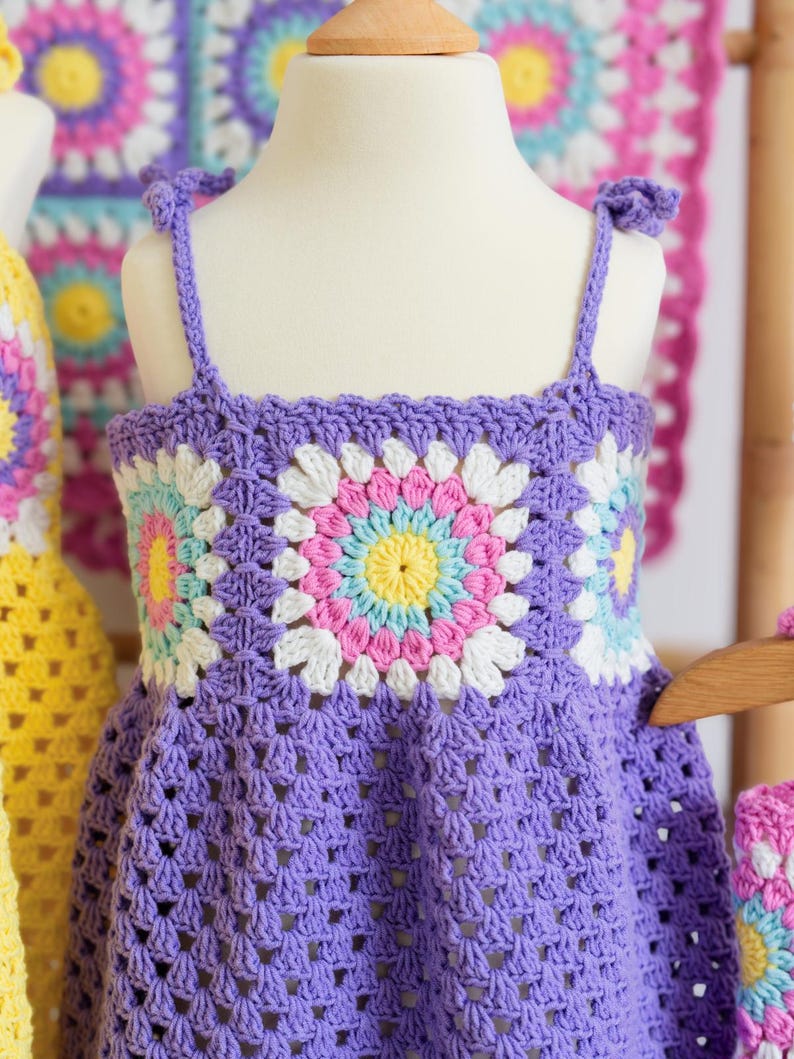 May include: A lilac-coloured, handmade crochet dress for a child, shown on a mannequin. The dress has a floral pattern on the bodice with white, pink, yellow, and blue details. The straps are tied at the shoulders.