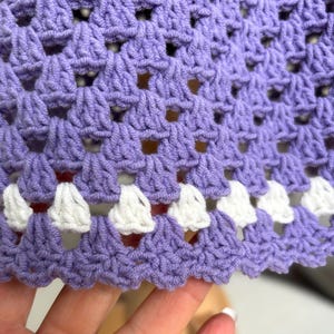 May include: Close-up of a crocheted fabric in a light purple hue. The crochet pattern features openwork squares and a scalloped edge with white accents. The texture of the yarn is visible, creating a tactile appearance.