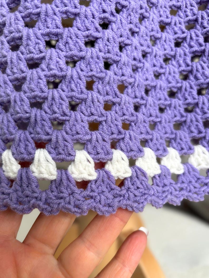 May include: Close-up of a crocheted fabric in a light purple hue. The crochet pattern features openwork squares and a scalloped edge with white accents. The texture of the yarn is visible, creating a tactile appearance.