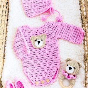 May include: Pink crocheted baby outfit with a bear applique, matching hat, booties, and a crocheted bear rattle.