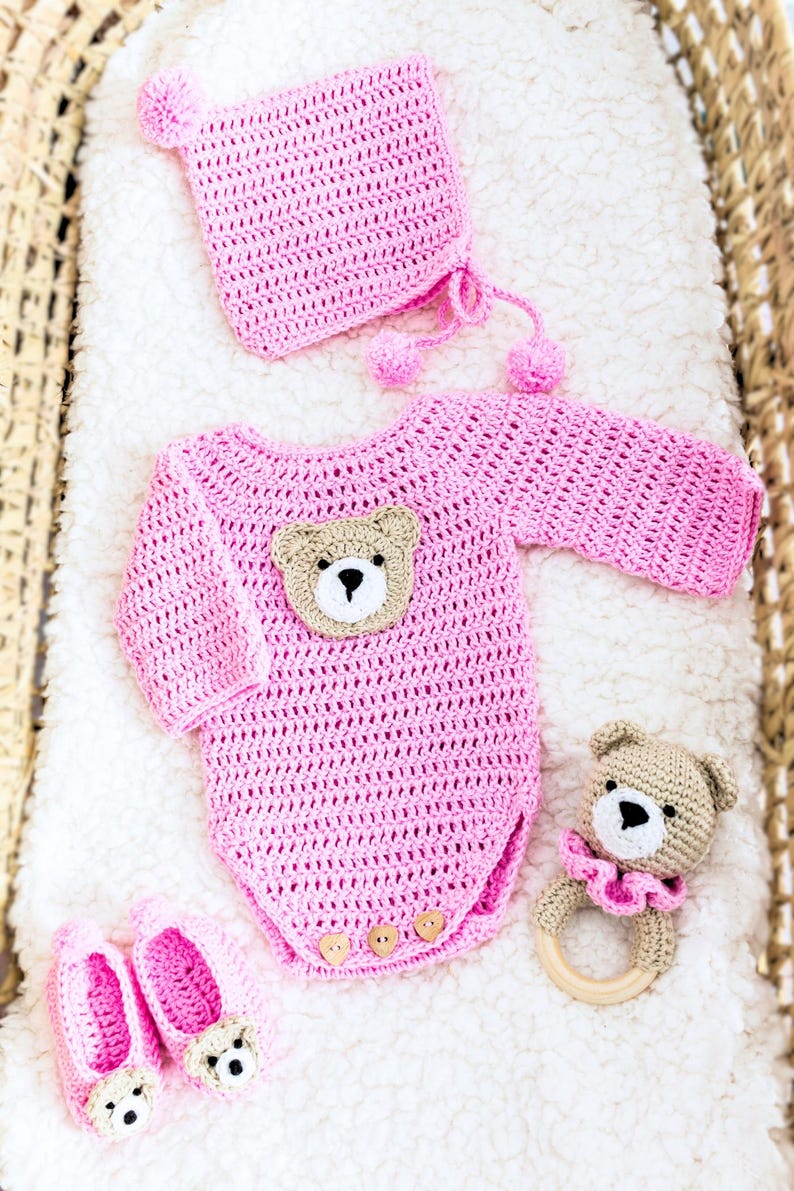 May include: Pink crocheted baby outfit with a bear applique, matching hat, booties, and a crocheted bear rattle.