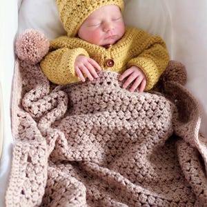 May include: A mustard yellow crocheted baby hat with a brown faux fur pom-pom, a matching mustard yellow crocheted long-sleeve cardigan with a single brown button, and a textured light beige crocheted baby blanket with a matching beige pom-pom.