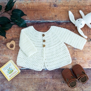 May include: A cream-colored long-sleeve crochet cardigan featuring three small brown flower-shaped buttons down the center front. The garment has a round neckline and a textured stitch pattern throughout the body and sleeves.