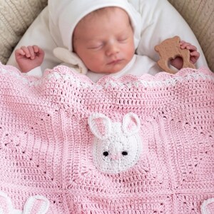 A baby asleep under a pink crocheted blanket featuring white bunny designs. The blanket is in a cream-coloured bassinet. The baby wears a white hat, and a wooden toy is present. The blanket is handmade.