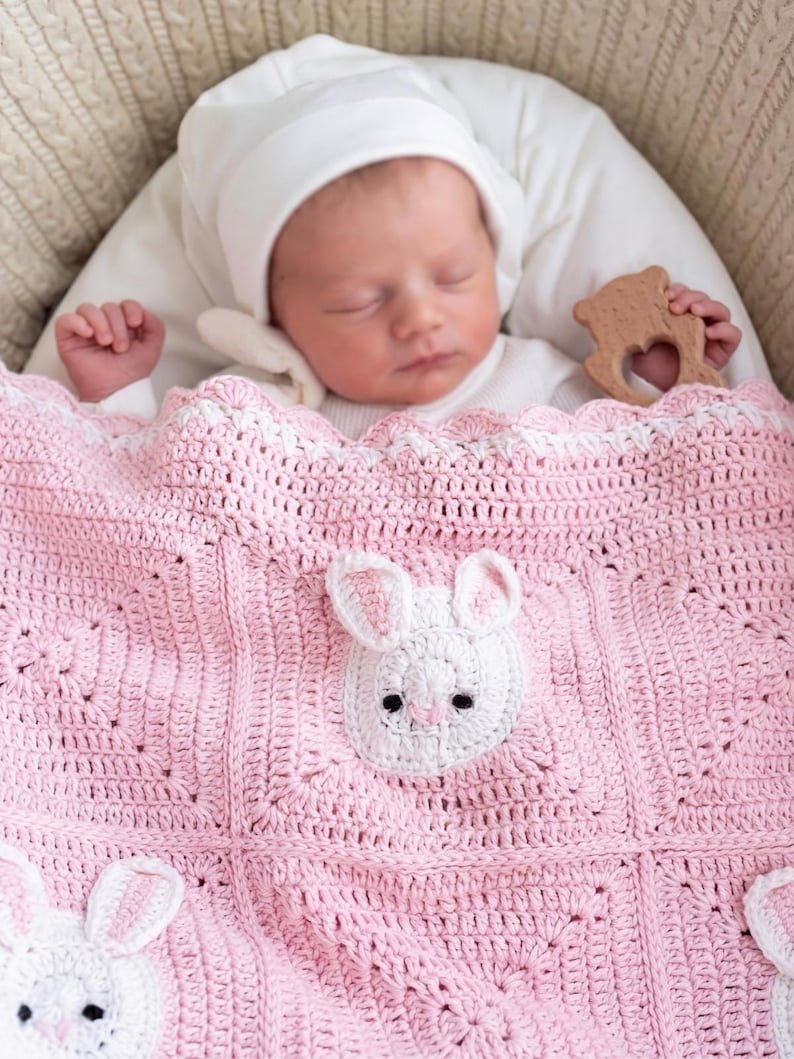 A baby asleep under a pink crocheted blanket featuring white bunny designs. The blanket is in a cream-coloured bassinet. The baby wears a white hat, and a wooden toy is present. The blanket is handmade.