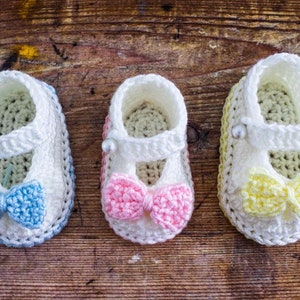 May include: Three pairs of crocheted baby booties with different colored bows. The booties are white with a white crocheted strap and a pearl embellishment. The bows are blue, pink, and yellow.