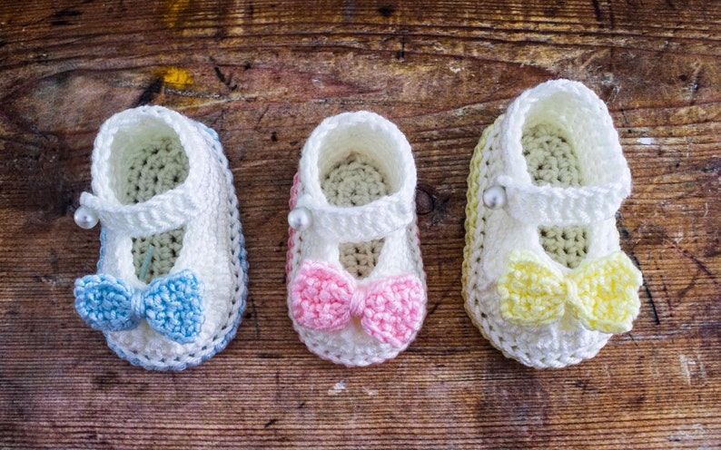 May include: Three pairs of crocheted baby booties with different colored bows. The booties are white with a white crocheted strap and a pearl embellishment. The bows are blue, pink, and yellow.
