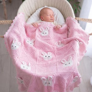 A baby asleep in a white wicker bassinet, covered by a pink crocheted blanket. The blanket features white bunny appliqués and a scalloped edge. The baby wears a white hat. The blanket is made of soft yarn.