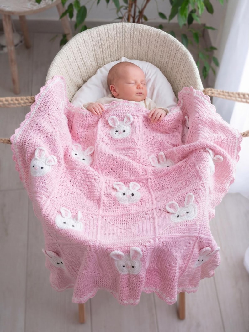 A baby asleep in a white wicker bassinet, covered by a pink crocheted blanket. The blanket features white bunny appliqués and a scalloped edge. The baby wears a white hat. The blanket is made of soft yarn.