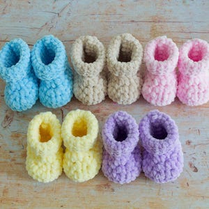 May include: A collection of eight soft, chunky knit booties arranged in two rows. The colors include light blue, beige, light pink, pale yellow, and lavender. Each pair features a thick, textured yarn construction with a rounded toe and cuff.