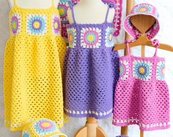 May include: A display of handmade crochet dresses and hats in yellow, purple, and pink. Each dress features a square pattern with colorful floral designs. A matching blanket is also displayed, showcasing the same pattern.