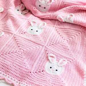 K&ouml;nnte beinhalten: Eine rosa geh&auml;kelte Babydecke mit wei&szlig;en Hasengesichtern, die auf der Oberfl&auml;che angebracht sind. Die Decke hat einen gewellten wei&szlig;en Rand und ist auf einer wei&szlig;en Oberfl&auml;che gefaltet. Die Hasengesichter haben schwarze Augen und rosa Innenohren.