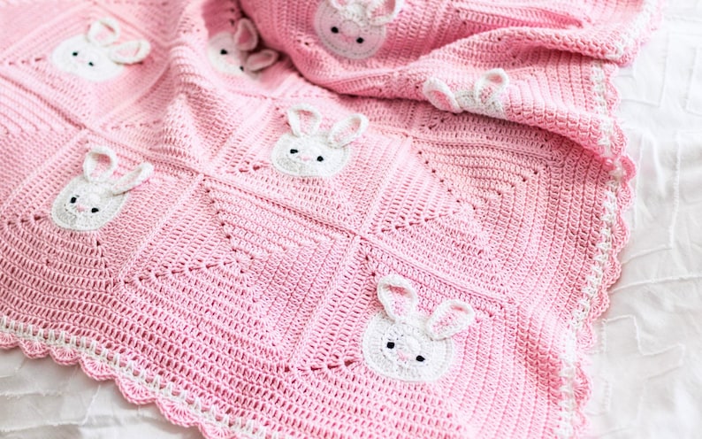 K&ouml;nnte beinhalten: Eine rosa geh&auml;kelte Babydecke mit wei&szlig;en Hasengesichtern, die auf der Oberfl&auml;che angebracht sind. Die Decke hat einen gewellten wei&szlig;en Rand und ist auf einer wei&szlig;en Oberfl&auml;che gefaltet. Die Hasengesichter haben schwarze Augen und rosa Innenohren.