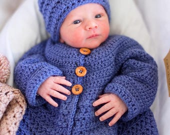 May include: A blue crocheted long-sleeve cardigan with three light brown wooden buttons down the front, paired with a matching blue crocheted beanie hat featuring a soft, fluffy, light pink pom-pom on the top of the crown.