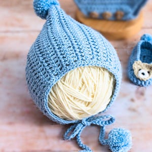 May include: A light blue crocheted baby hat with a pom-pom on top and a white yarn ball inside. The hat has two ties with pom-poms on the ends.