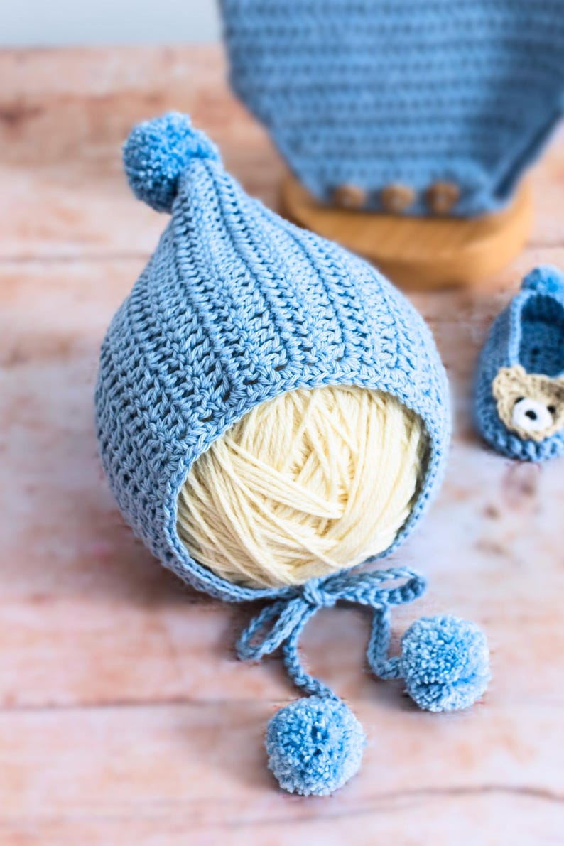 May include: A light blue crocheted baby hat with a pom-pom on top and a white yarn ball inside. The hat has two ties with pom-poms on the ends.