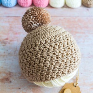 May include: A brown crocheted baby hat with a brown pom-pom on top. The hat is sitting on a wooden surface with a brown paper tag with a heart cutout.
