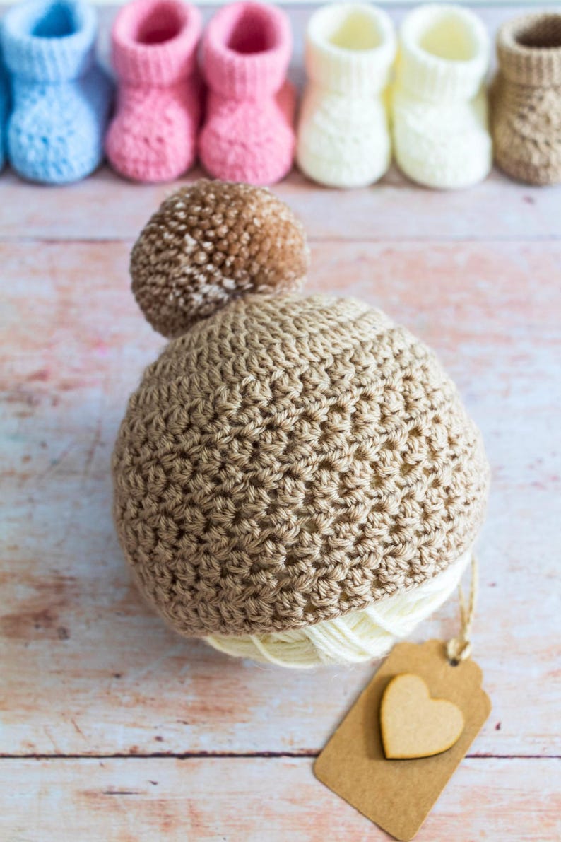 May include: A brown crocheted baby hat with a brown pom-pom on top. The hat is sitting on a wooden surface with a brown paper tag with a heart cutout.