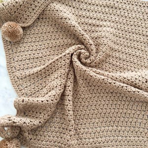 May include: A light brown crochet blanket featuring a textured stitch pattern and decorative pom-poms attached to the corners. The fabric is folded in the center, showcasing the intricate yarn work and the consistent tan color throughout the piece.