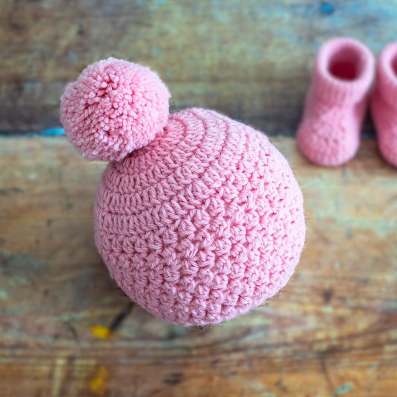 May include: A pink crocheted baby hat with a pom-pom on top. The hat is lying on a wooden surface.