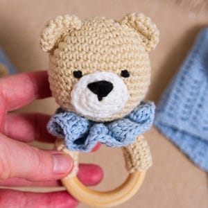 K&ouml;nnte beinhalten: Ein geh&auml;keltes Teddyb&auml;r-Rassel mit einem blauen Kragen und einem Holzring. Die Rassel wird in einer Hand gehalten.