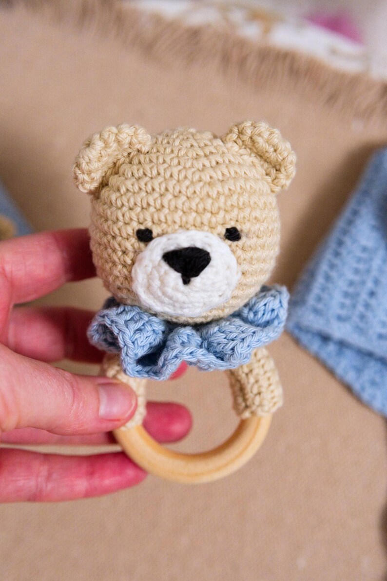 K&ouml;nnte beinhalten: Ein geh&auml;keltes Teddyb&auml;r-Rassel mit einem blauen Kragen und einem Holzring. Die Rassel wird in einer Hand gehalten.