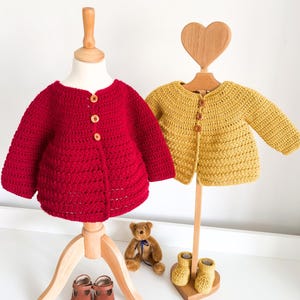 May include: A red crocheted cardigan with three wooden buttons and a yellow crocheted cardigan with three wooden buttons, displayed on two wooden stands. A small brown teddy bear sits between the stands, and a pair of brown leather shoes are placed near the red cardigan, while yellow crocheted booties sit near the yellow cardigan.