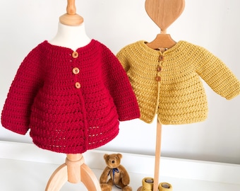 Könnte beinhalten: Eine rote gehäkelte Strickjacke mit drei Holzknöpfen und eine gelbe gehäkelte Strickjacke mit drei Holzknöpfen, präsentiert auf zwei Holzständern. Ein kleiner brauner Teddybär sitzt zwischen den Ständern, ein Paar braune Lederschuhe steht neben der roten Strickjacke und gelbe gehäkelte Babyschuhe stehen neben der gelben Strickjacke.