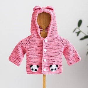May include: A pink crocheted baby cardigan with a hood and panda bear appliques on the front pockets. The cardigan has white buttons and is displayed on a wooden stand.