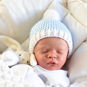 May include: A sleeping baby wearing a blue and white crocheted hat with a white pom-pom on top. The baby is lying on a white blanket with a white stuffed bunny.