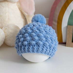 May include: A light blue crocheted baby hat with a pom pom on top. The hat is made with a textured stitch that creates a bumpy surface.