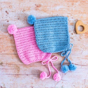 Könnte beinhalten: Zwei gehäkelte Baby-Mützen mit Bommeln. Eine Mütze ist rosa und die andere blau.