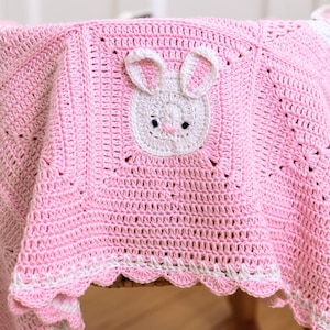 K&ouml;nnte beinhalten: Eine rosa geh&auml;kelte Babydecke mit wei&szlig;en Hasengesichtern und Ohren. Die Decke hat einen gewellten wei&szlig;en Rand und ist &uuml;ber einer Holzoberfl&auml;che drapiert. Die Hasengesichter sind auf der Decke und an den Ecken zentriert.