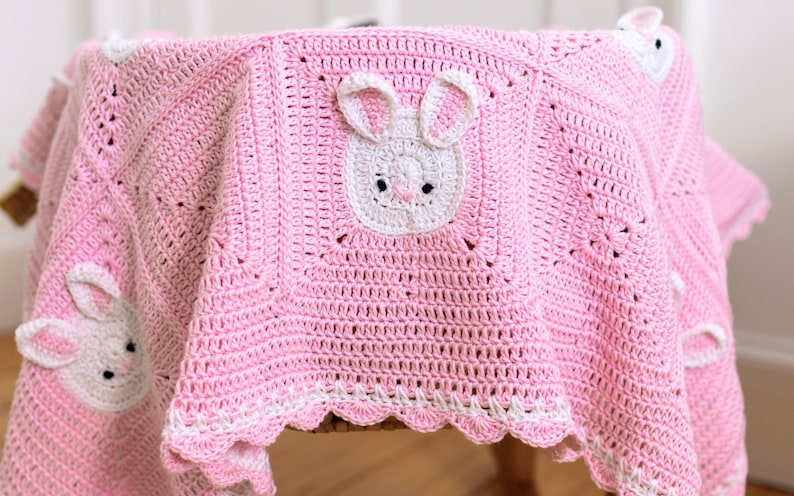 K&ouml;nnte beinhalten: Eine rosa geh&auml;kelte Babydecke mit wei&szlig;en Hasengesichtern und Ohren. Die Decke hat einen gewellten wei&szlig;en Rand und ist &uuml;ber einer Holzoberfl&auml;che drapiert. Die Hasengesichter sind auf der Decke und an den Ecken zentriert.