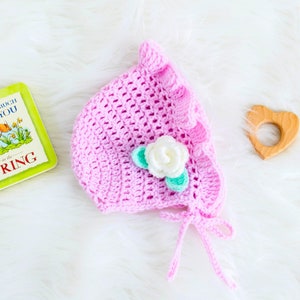 May include: A pink crocheted baby bonnet with a white flower and green leaves. The bonnet has a ruffle trim and ties at the back.
