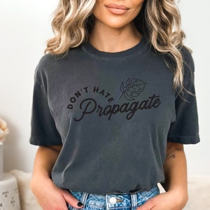 May include: A dark gray t-shirt with a black graphic that says "Don't Hate Propagate" with a small leaf design. The shirt is being worn by a person with long blonde hair and blue denim shorts.