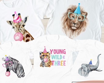 May include: Five white t-shirts featuring different animal designs with party hats. The designs include a giraffe, a lion, a zebra, a cheetah, and an elephant. Each animal is wearing a party hat and has a pink bubble in its mouth. One of the shirts has the text "Young Wild & Three" on it.