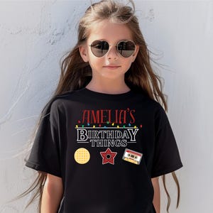 May include: Black t-shirt with the text "AMELIA'S BIRTHDAY THINGS" in red and white. The shirt features a waffle, a red star, and a cassette tape graphic. The shirt is worn by a young person wearing heart-shaped sunglasses.