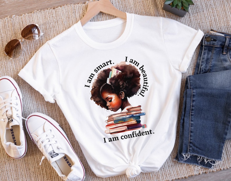 May include: White t-shirt with a graphic of a young Black girl with a stack of books on her head. The text on the shirt reads "I am smart. I am beautiful. I am confident."