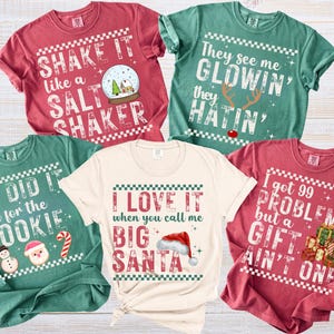 Op de afbeelding: Een verzameling van vijf t-shirts met korte mouwen in verschillende kleuren, elk met een kerstthema en tekst. De shirts zijn rood, groen en cr&egrave;mekleurig, met zinnen als "Shake it like a salt shaker" en "I love it when you call me Big Santa".